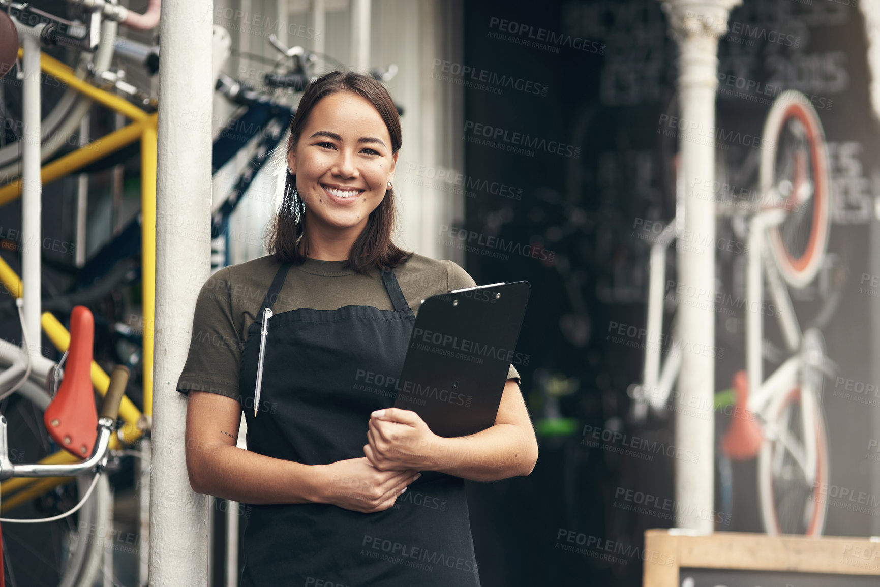 Buy stock photo Shot of an attractive young woman standing alone outside her bicycle shop and holding a clipboard