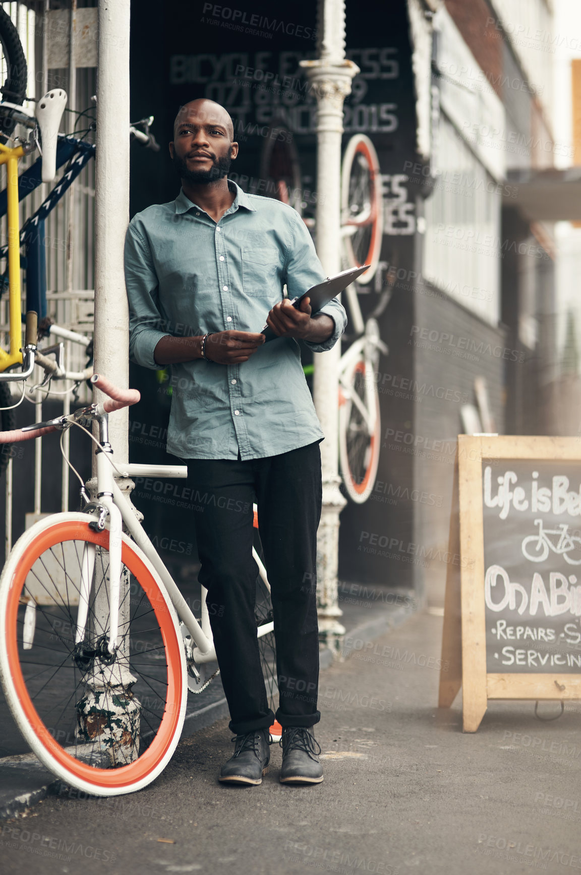 Buy stock photo Full length shot of a handsome young man standing outside his bicycle shop and looking contemplative while holding a clipboard