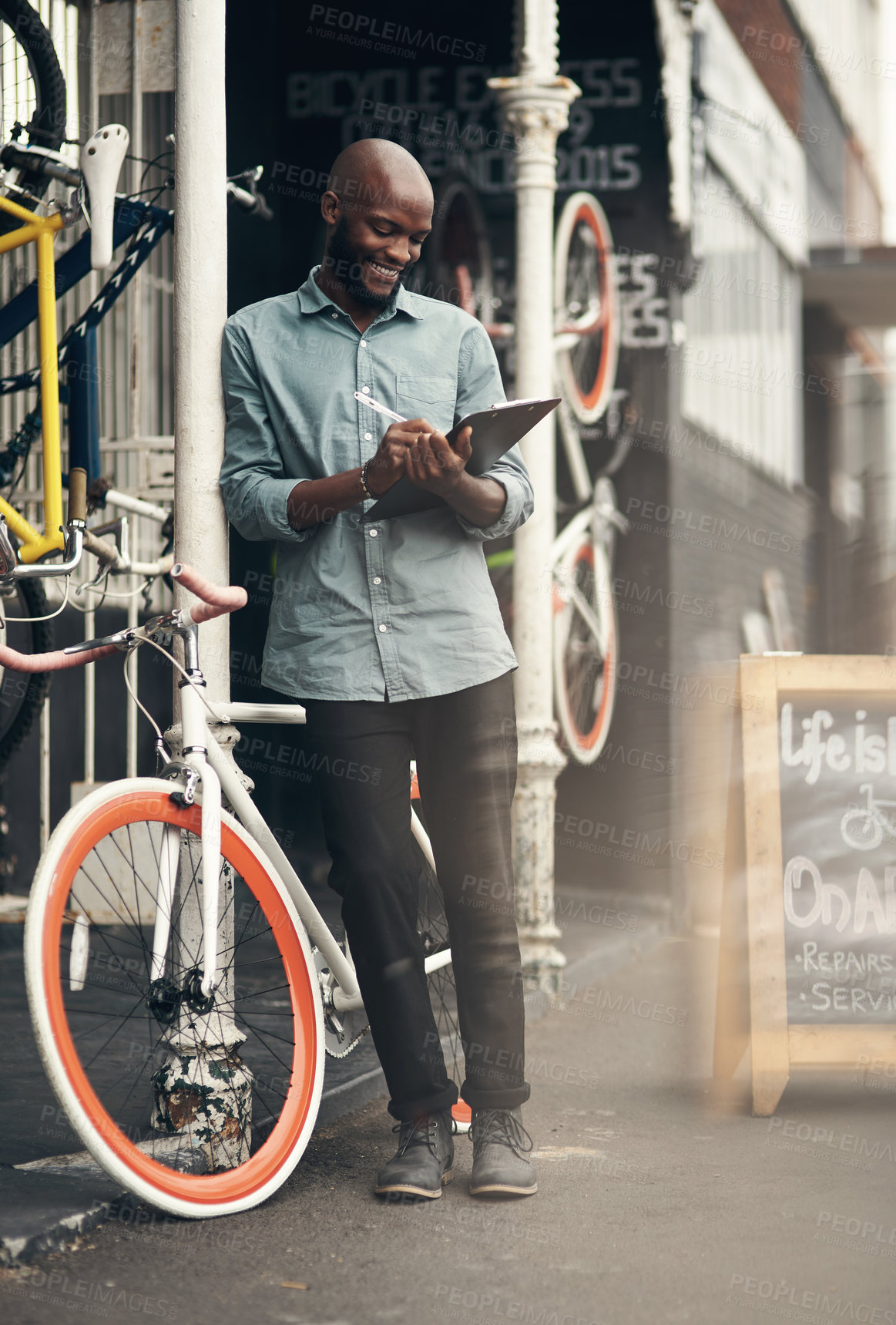 Buy stock photo Full length shot of a handsome young man standing alone outside his bicycle shop and writing on a clipboard