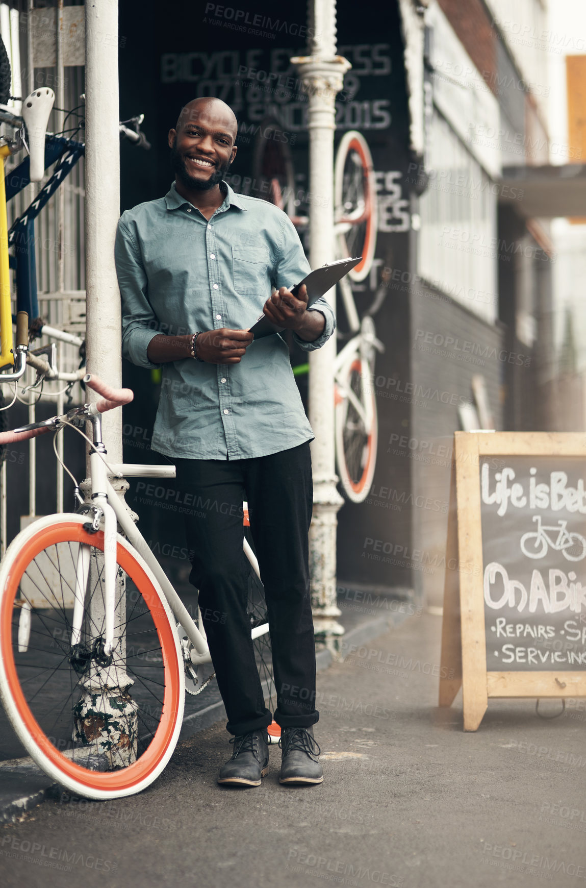 Buy stock photo Full length shot of a handsome young man standing alone outside his bicycle shop and holding a clipboard