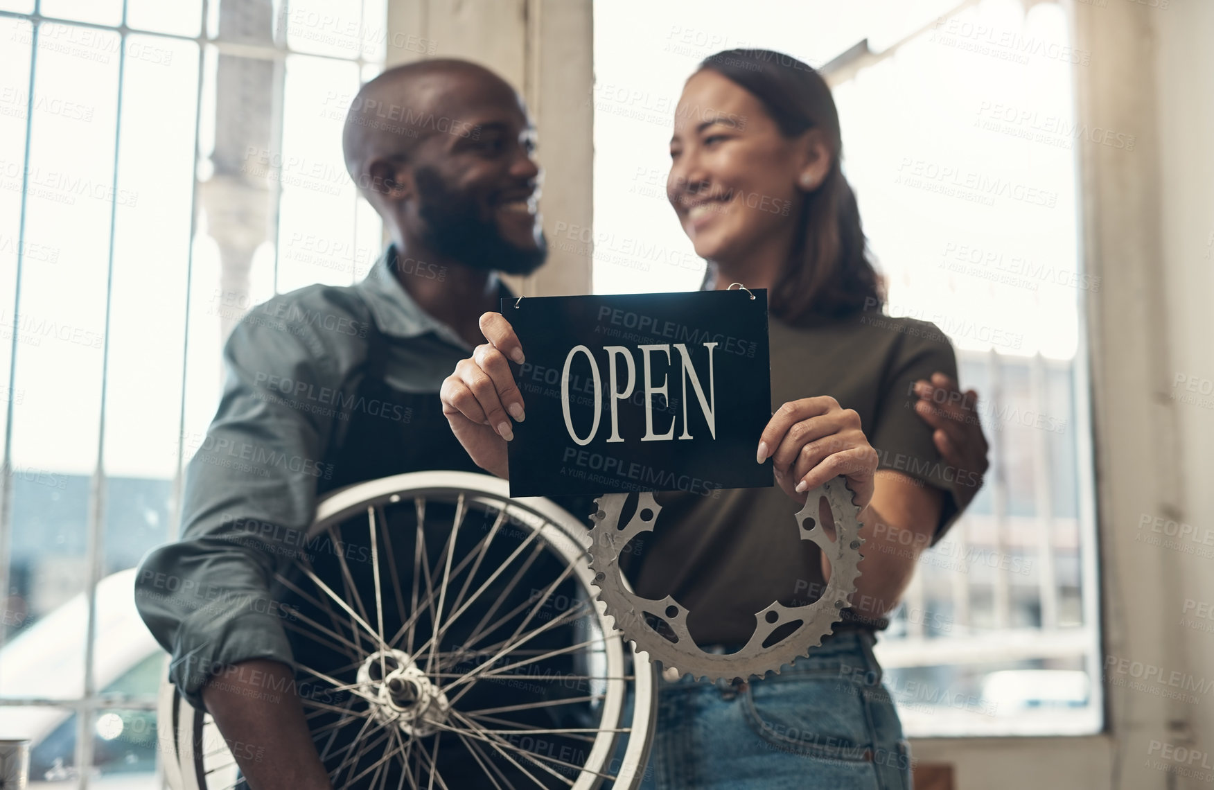 Buy stock photo Shot of a young couple standing together in their bicycle shop and holding an open sign