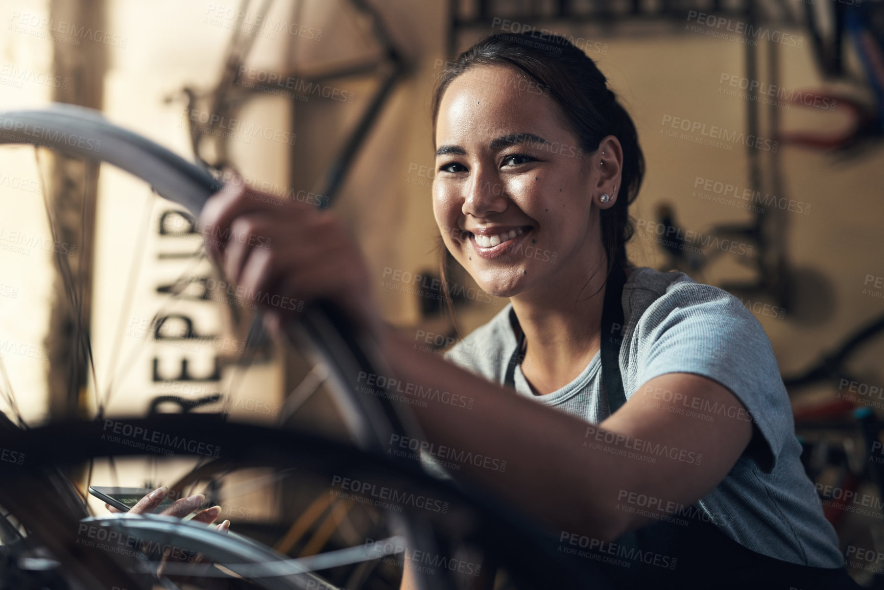 Buy stock photo Portrait of a young woman looking extremely pleased while holding a cellphone and fixing a bike at a bicycle repair shop