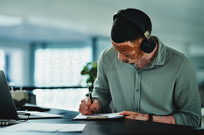 Buy stock photo Shot of a young businessman doing paperwork while wearing headphones in an office at work