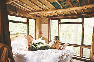 Buy stock photo Shot of a young woman reading a book while lying on a bed in a cabin