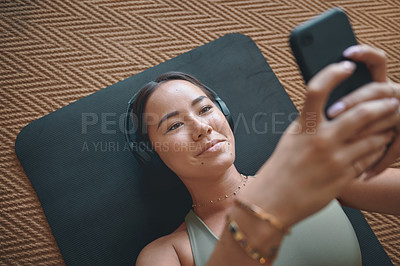 Buy stock photo High angle shot of a young woman wearing headphones and using a cellphone while lying on an exercise mat at home