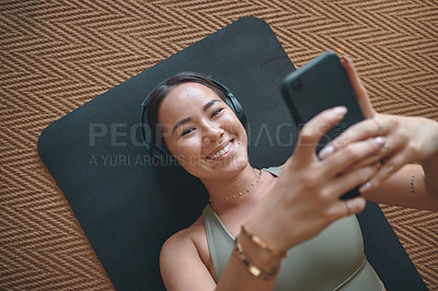 Buy stock photo Portrait of a young woman wearing headphones and using a cellphone while lying on an exercise mat at home
