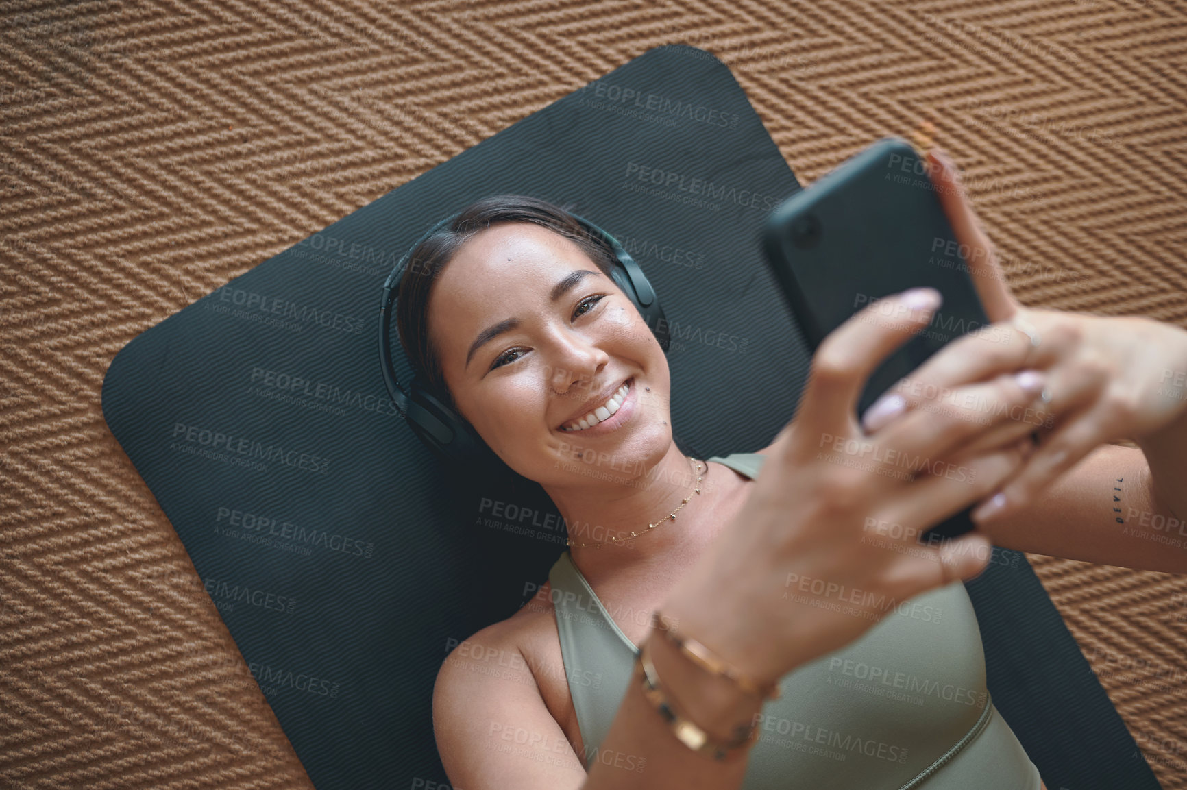 Buy stock photo Portrait of a young woman wearing headphones and using a cellphone while lying on an exercise mat at home