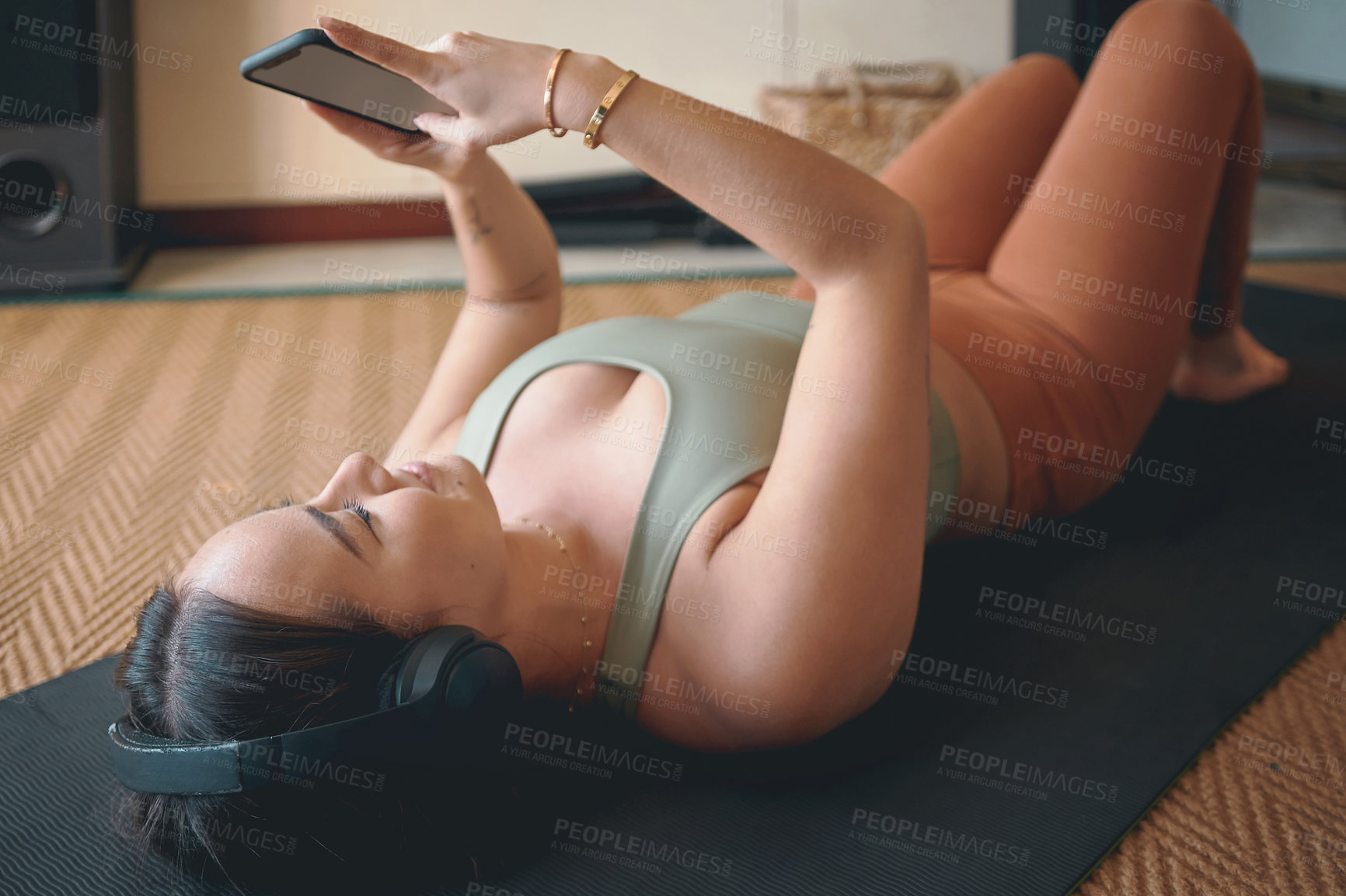 Buy stock photo Shot of a young woman wearing headphones and using a cellphone while lying on an exercise mat at home