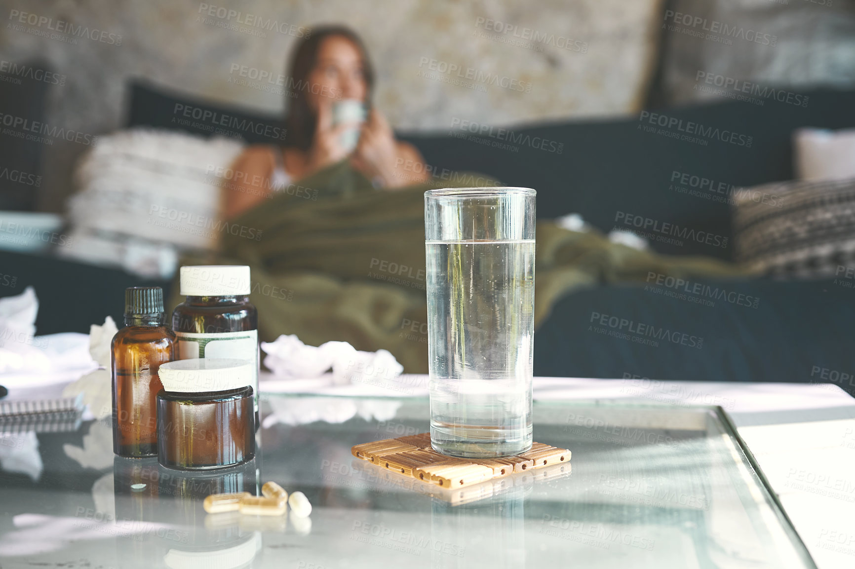 Buy stock photo Shot of a medication on a table with a woman recovering from an illness on the sofa in the background at home