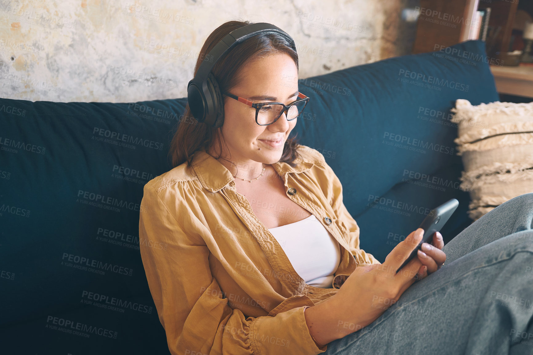 Buy stock photo Shot of a young woman using headphones and a smartphone while relaxing on the sofa at home