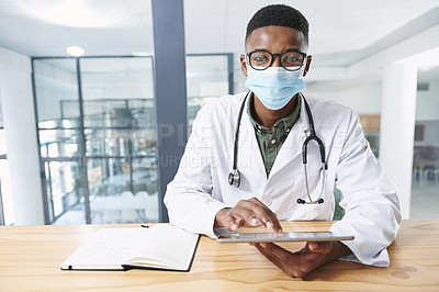 Buy stock photo Shot of a young doctor wearing a face mask and sitting in his office at the clinic while using technology