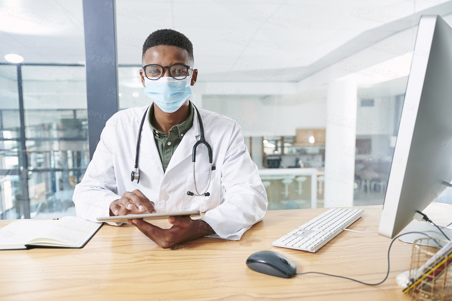 Buy stock photo Shot of a young doctor wearing a face mask and sitting in his office at the clinic while using technology