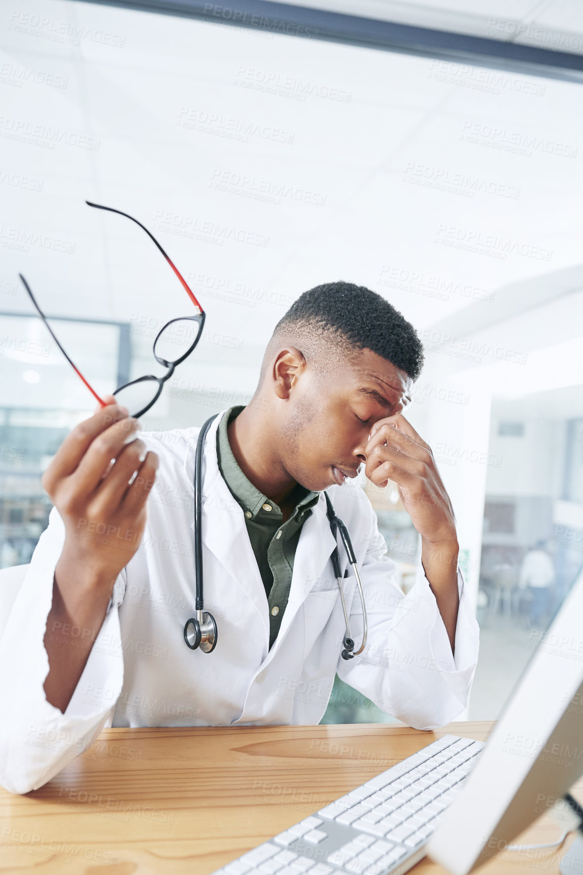 Buy stock photo Shot of a handsome young doctor sitting alone in his office at the clinic and feeling stressed using his computer