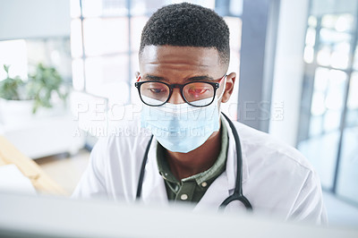 Buy stock photo Shot of a young doctor wearing a mask and sitting in his office at the clinic while using a computer