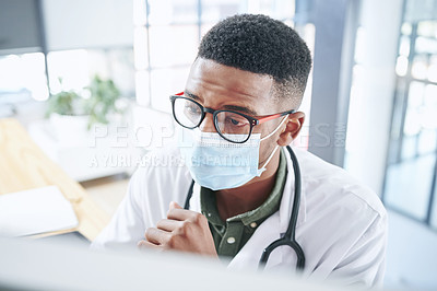 Buy stock photo Shot of a young doctor wearing a mask and sitting in his office at the clinic while using a computer