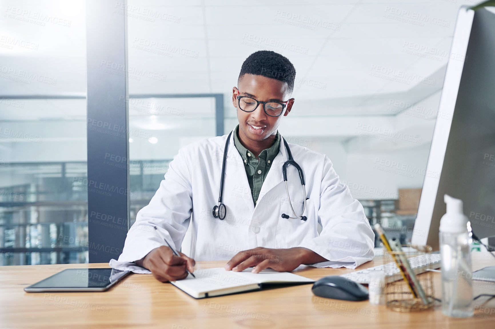 Buy stock photo Shot of a handsome young doctor sitting alone in his office at the clinic and writing in his notebook