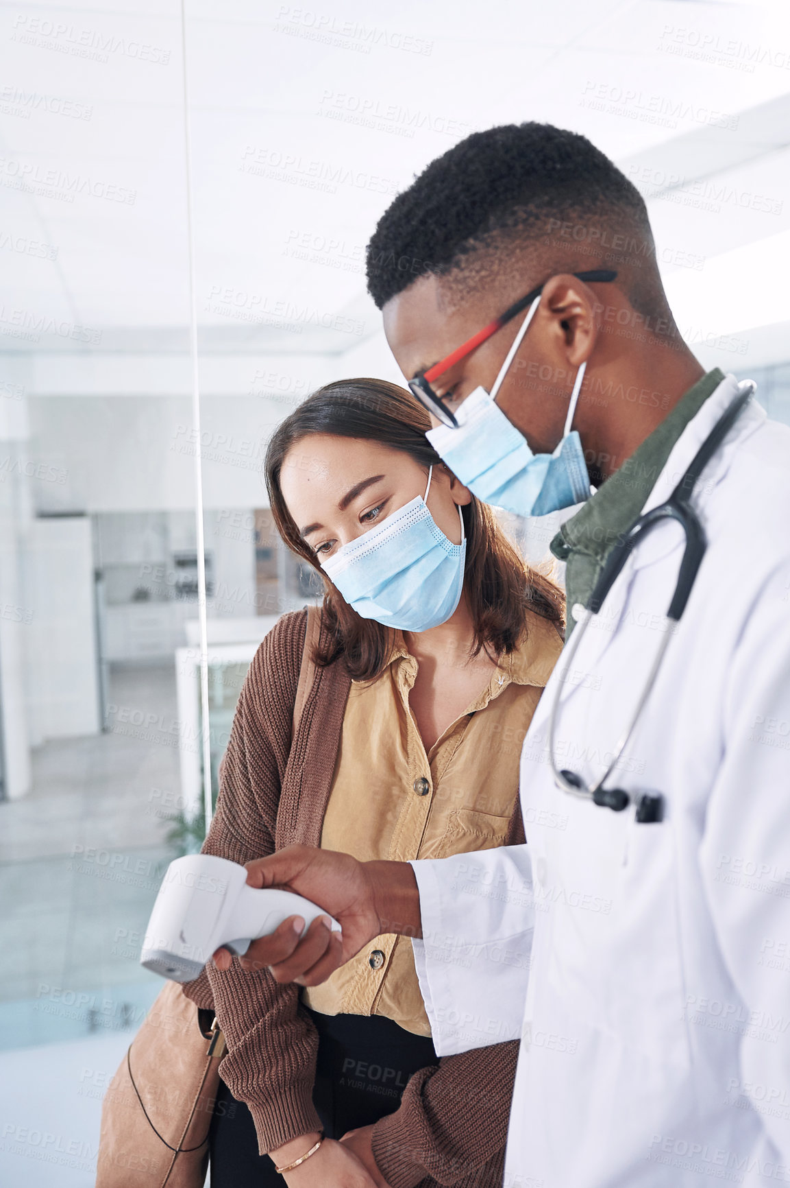 Buy stock photo Shot of a young doctor wearing a mask and standing with a patient in the clinic while taking her temperature