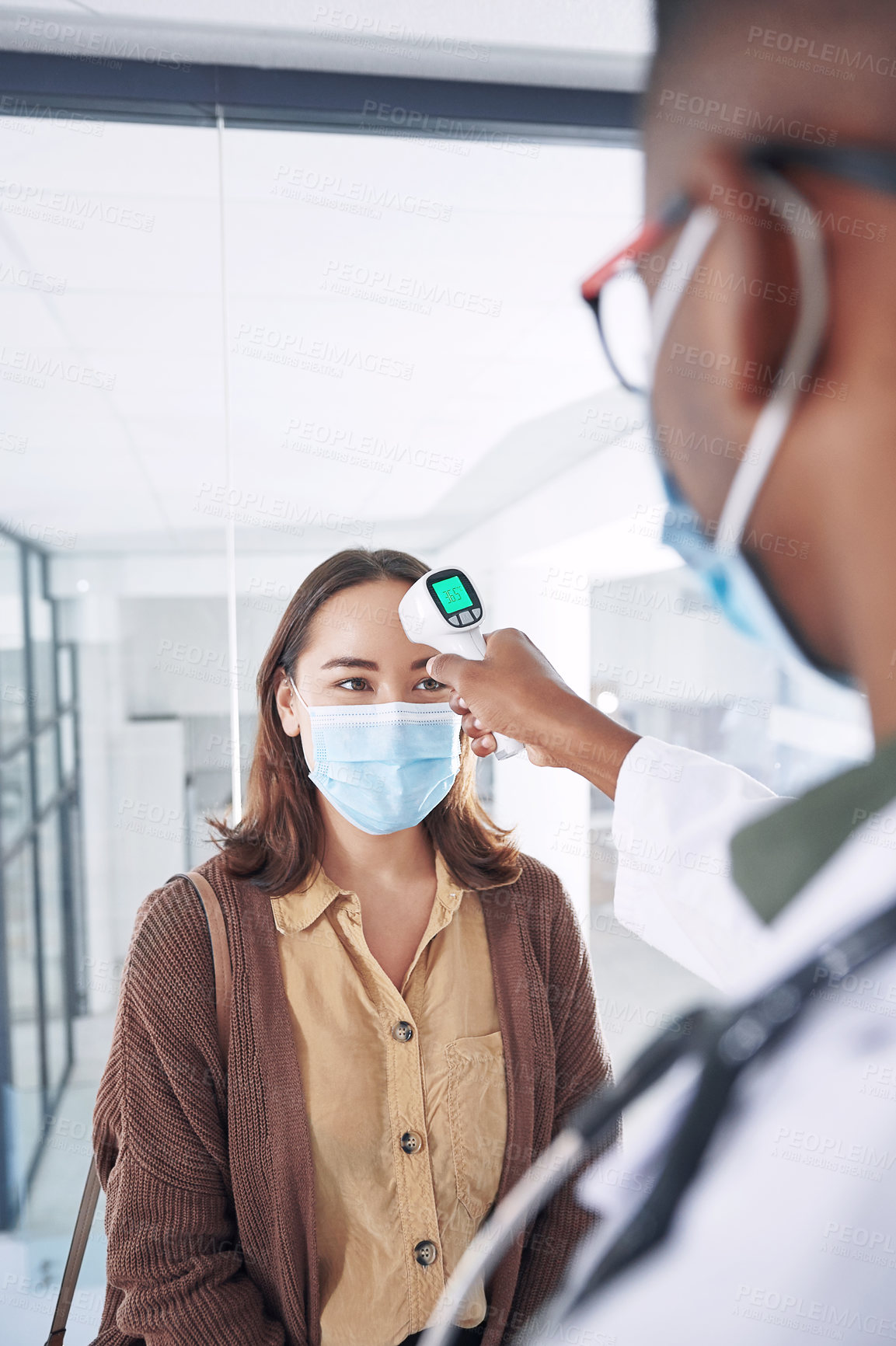 Buy stock photo Shot of a young woman wearing a mask and getting her temperature checked by her doctor in the clinic