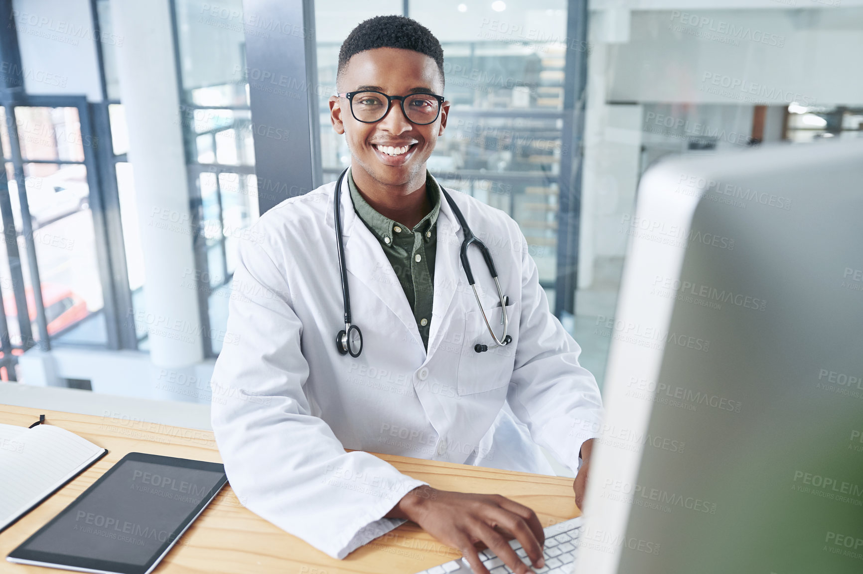 Buy stock photo Shot of a handsome young doctor sitting alone in his office at the clinic and using his computer