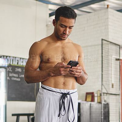 Buy stock photo Cropped shot of a handsome young man standing using his phone while standing shirtless in his jiu jitsu pants in the gym
