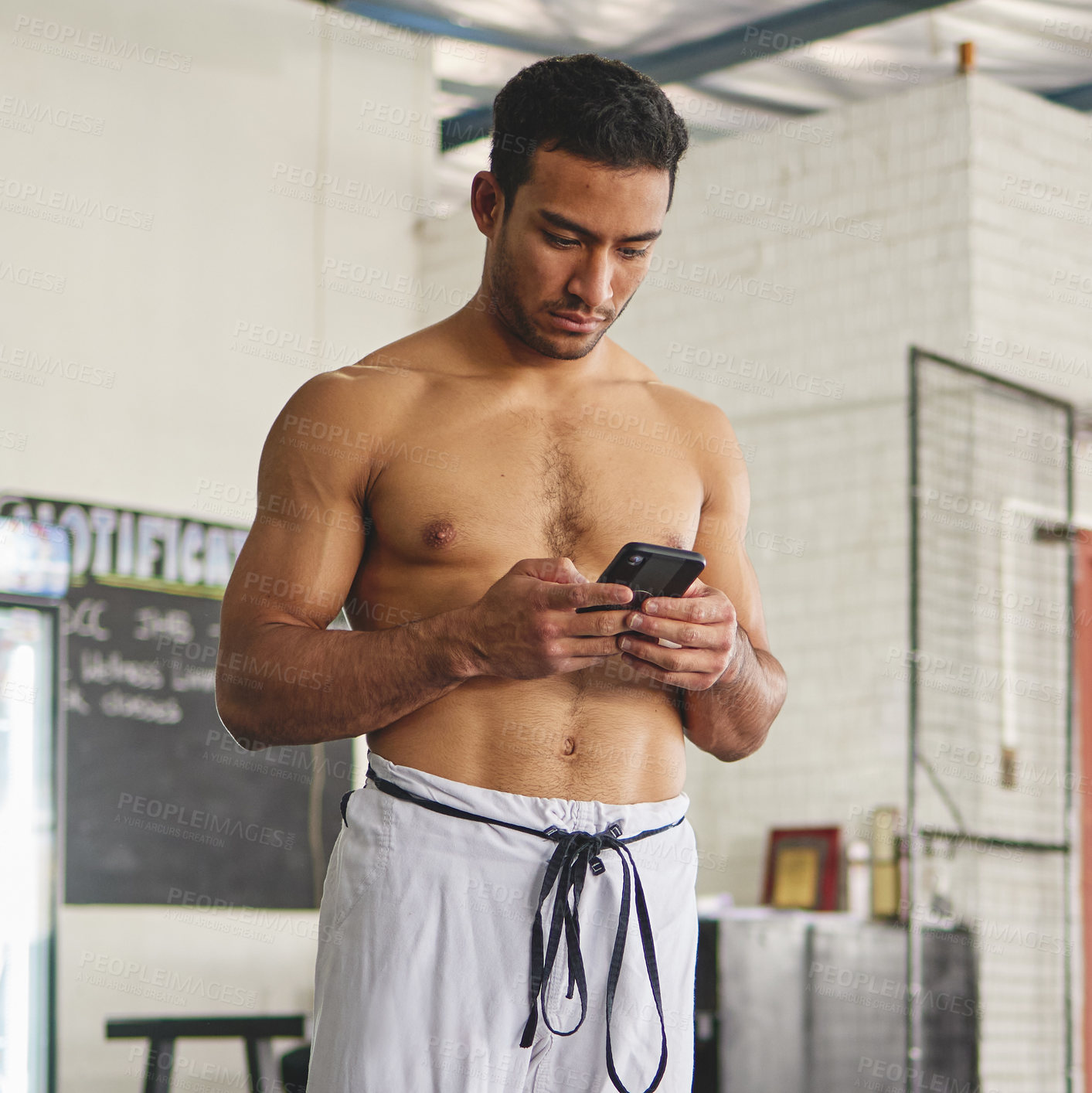 Buy stock photo Cropped shot of a handsome young man standing using his phone while standing shirtless in his jiu jitsu pants in the gym