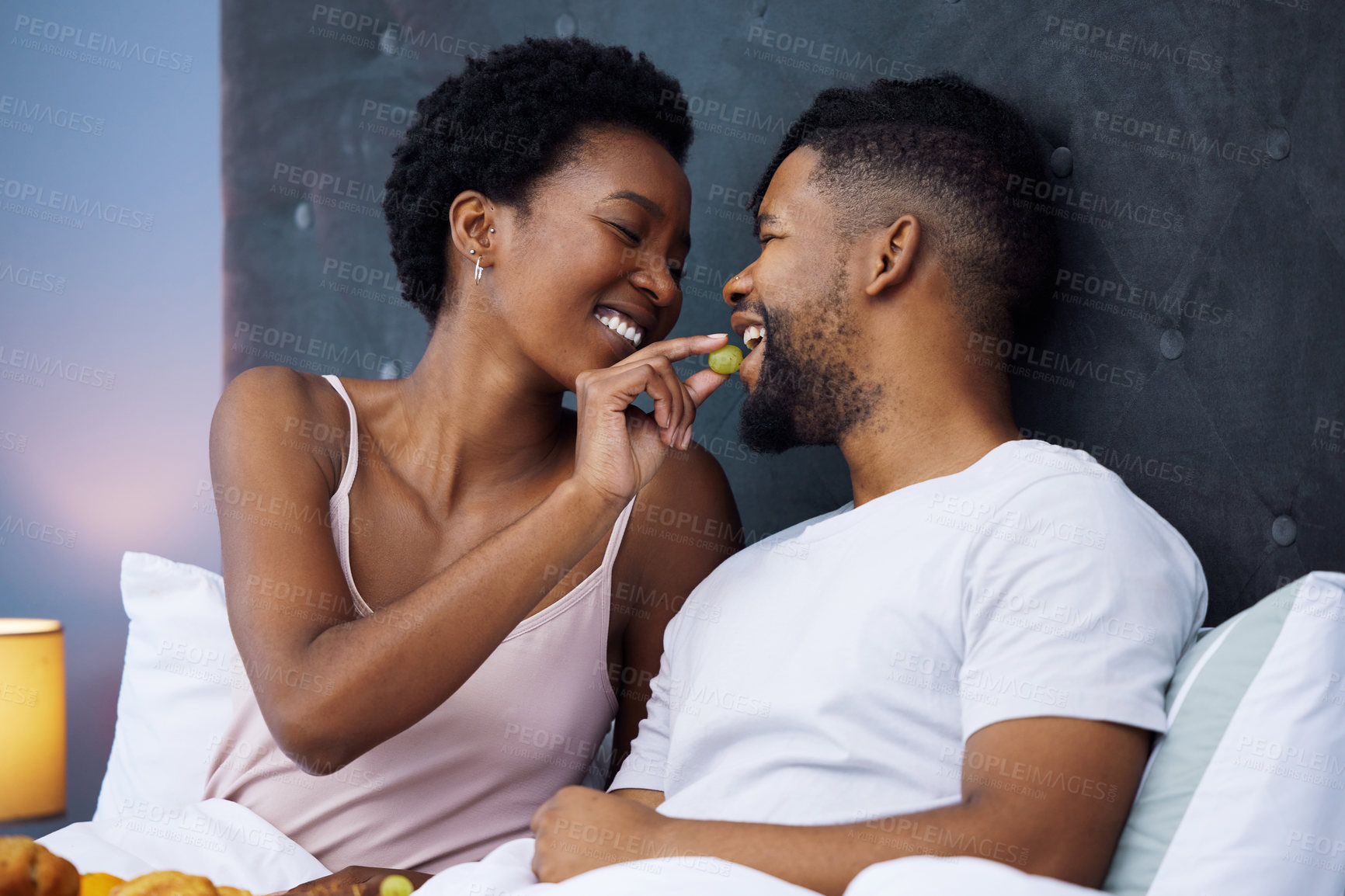 Buy stock photo Happy, couple and intimate in eating grapes in bed for romance, love or connection as partner in relationship. African woman and man for support with joyful, emotion and marriage for bonding or trust