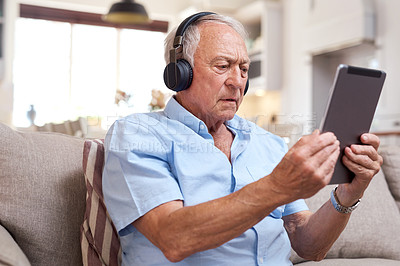 Buy stock photo Shot of an elderly man wearing headphones and using a digital tablet on the sofa at home