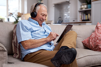 Buy stock photo Shot of an elderly man wearing headphones and using a digital tablet on the sofa at home