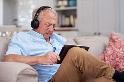 Buy stock photo Shot of an elderly man wearing headphones and using a digital tablet on the sofa at home