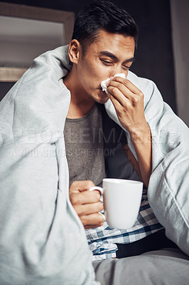 Buy stock photo Shot of a young man blowing his nose and having tea while recovering from an illness in bed at home