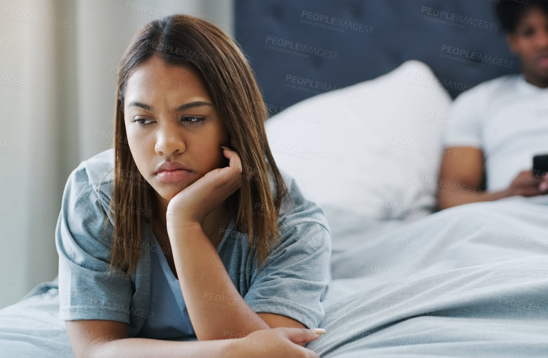 Buy stock photo Shot of a woman looking upset while lying on her bed with her boyfriend using his cellphone in the background