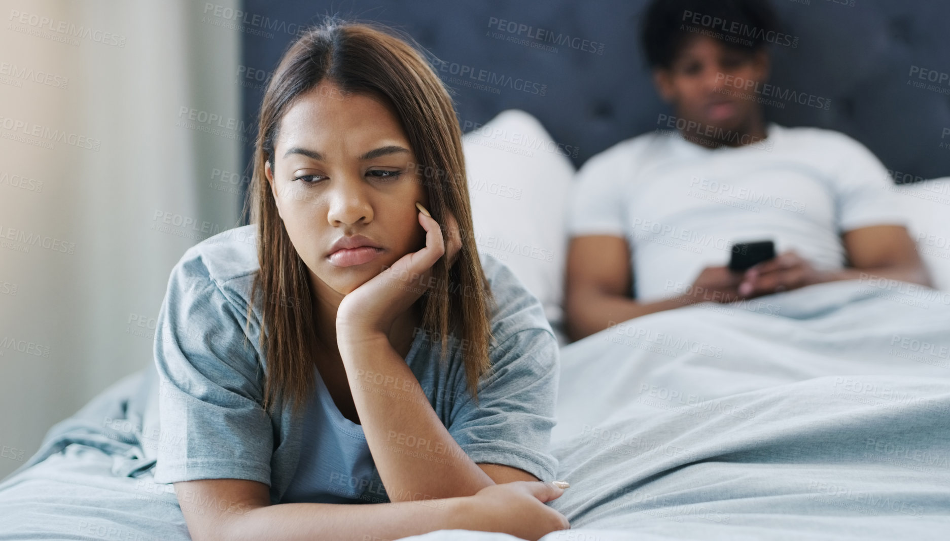 Buy stock photo Shot of a woman looking upset while lying on her bed with her boyfriend using his cellphone in the background