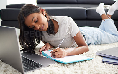 Buy stock photo Shot of a young woman lying on her living room floor and writing in a notebook while using her laptop