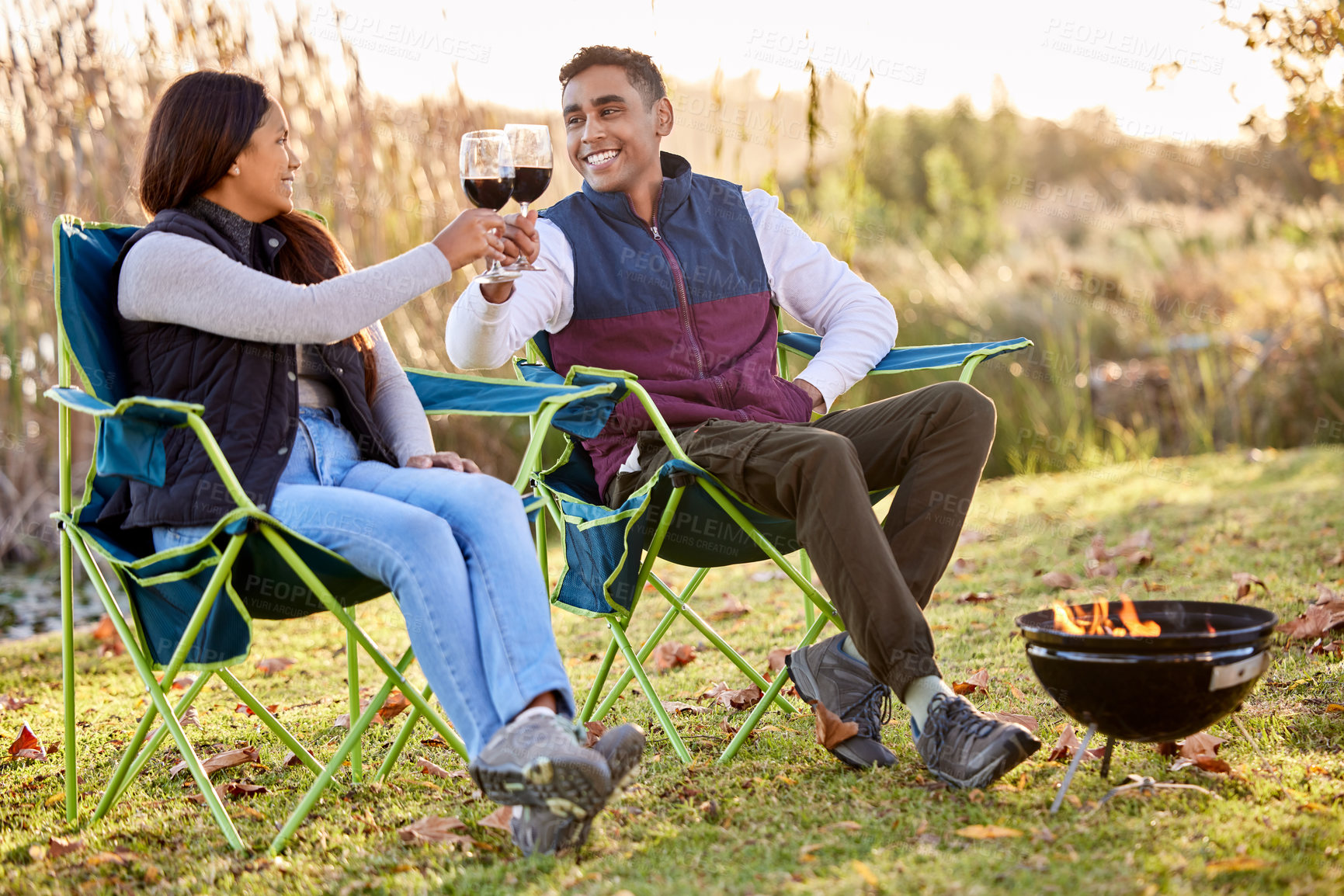 Buy stock photo Cheers, wine and happy couple camping in park for love, holiday vacation and valentines day picnic. Toast, man and woman with fireplace, smile and chairs on date for anniversary in nature or forest