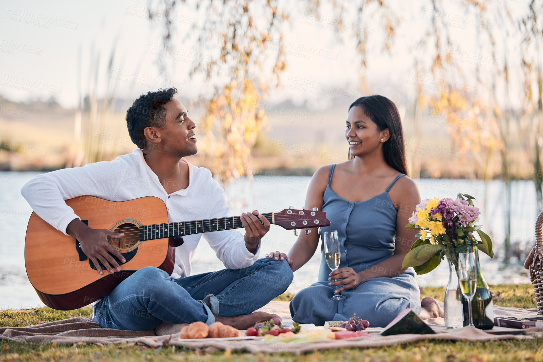 Buy stock photo Shot of a young man playing a guitar while on a picnic with his girlfriend at a lakeside