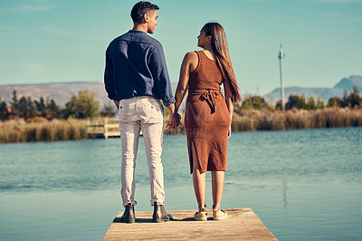 Buy stock photo Rearview shot of a young couple holding hands while standing together on a pier at a lakeside