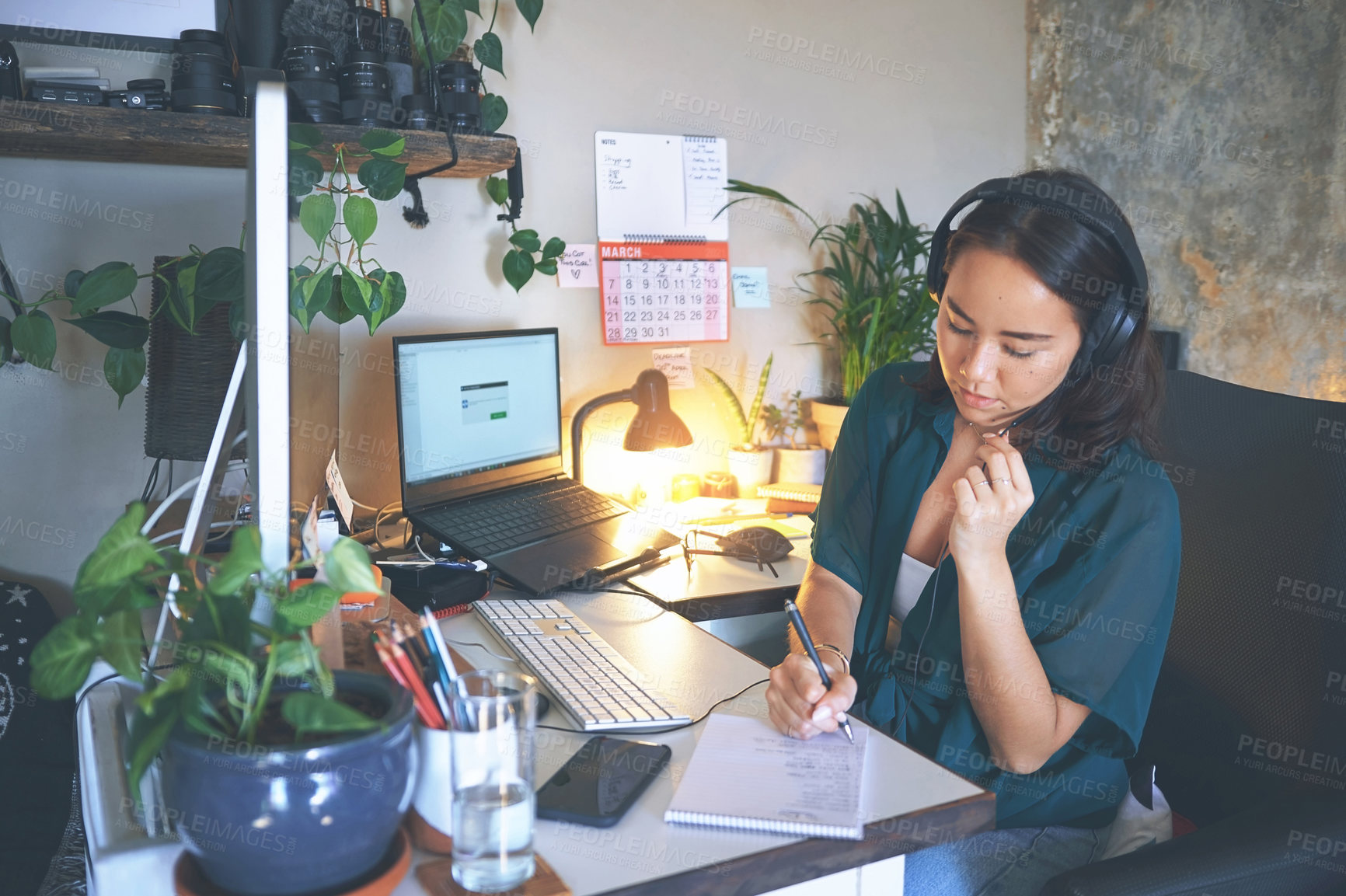 Buy stock photo Shot of an attractive young woman sitting alone and wearing a headset while writing notes in her home office