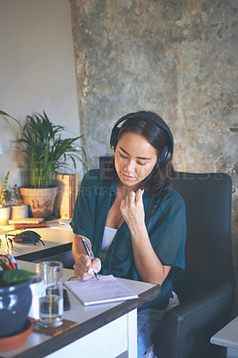 Buy stock photo Shot of an attractive young woman sitting alone and wearing a headset while writing notes in her home office