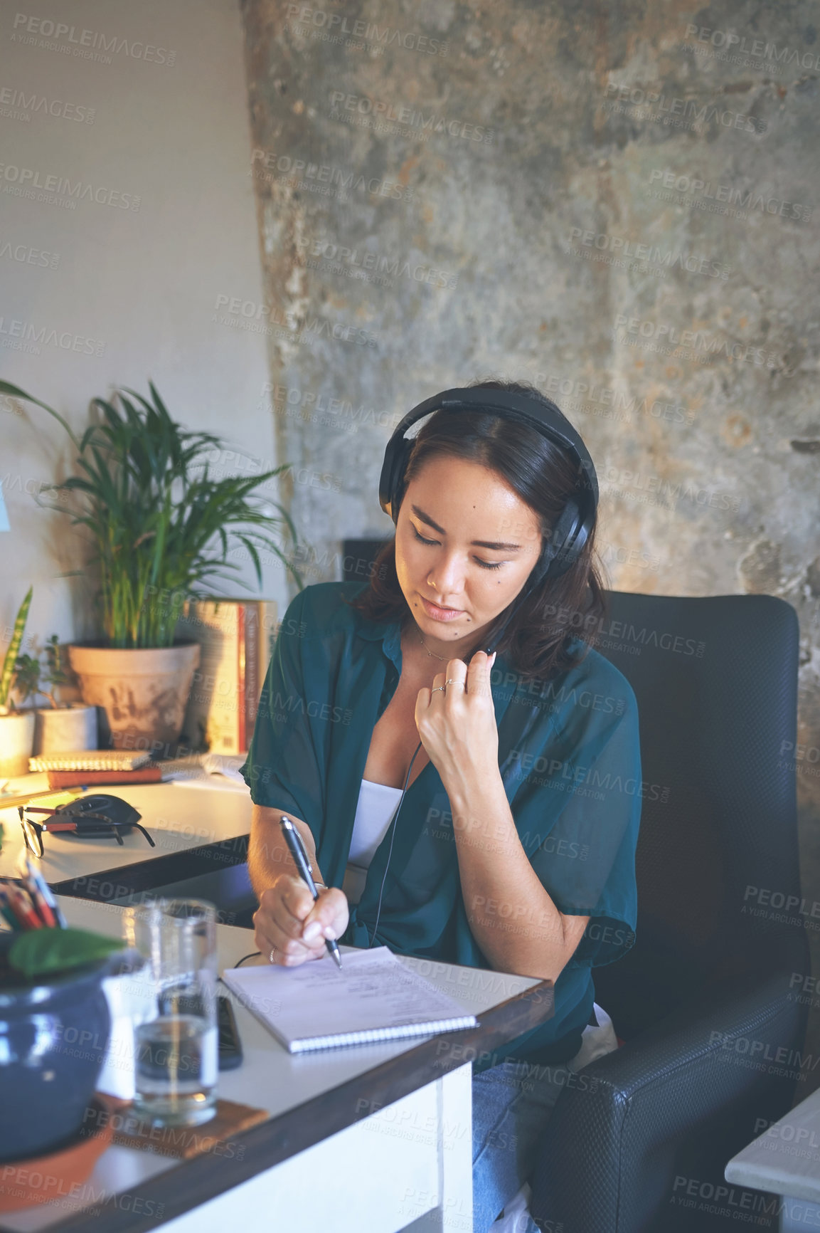 Buy stock photo Shot of an attractive young woman sitting alone and wearing a headset while writing notes in her home office