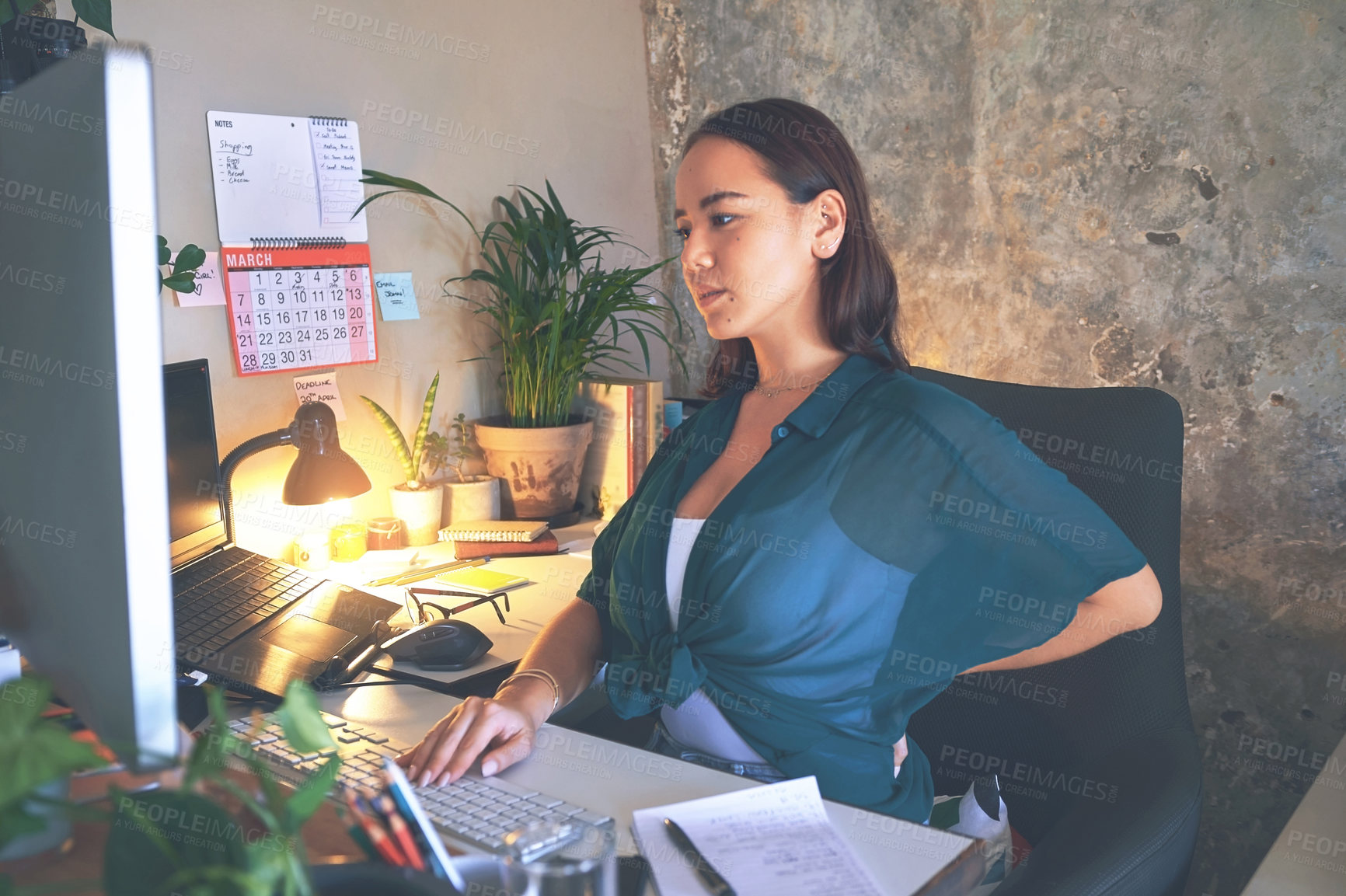 Buy stock photo Shot of a young woman sitting alone and suffering from back ache while using her computer to work from home