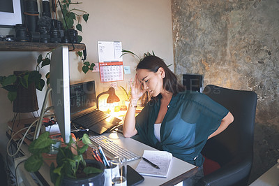 Buy stock photo Shot of a young woman sitting alone and suffering from back ache while using her computer to work from home