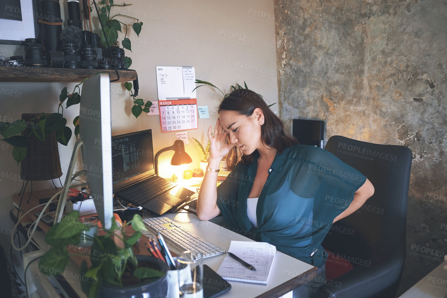 Buy stock photo Shot of a young woman sitting alone and suffering from back ache while using her computer to work from home