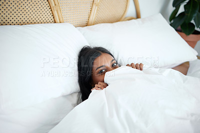 Buy stock photo Shot of a young woman peeking from under her duvet while lying in her bed