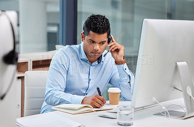 Buy stock photo Shot of a handsome young businessman sitting alone in his office and writing notes while wearing a headset