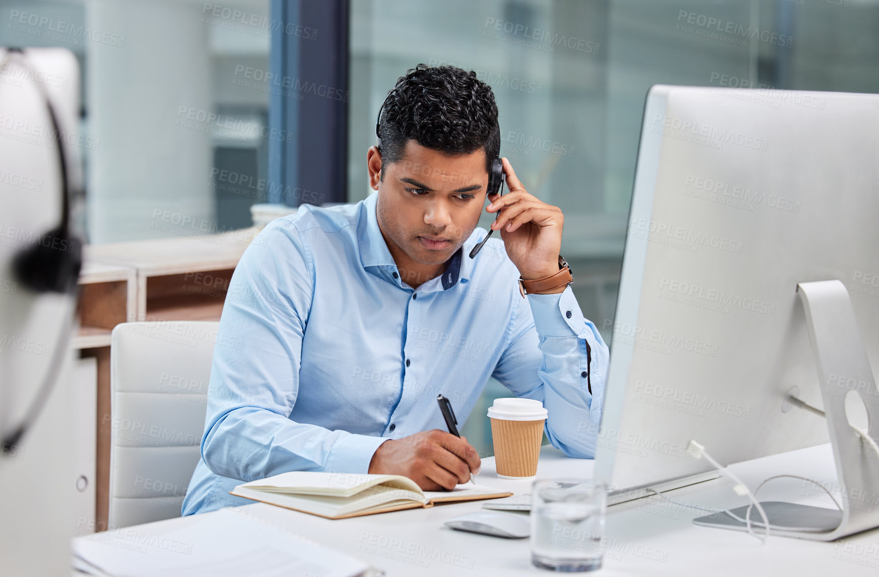 Buy stock photo Shot of a handsome young businessman sitting alone in his office and writing notes while wearing a headset