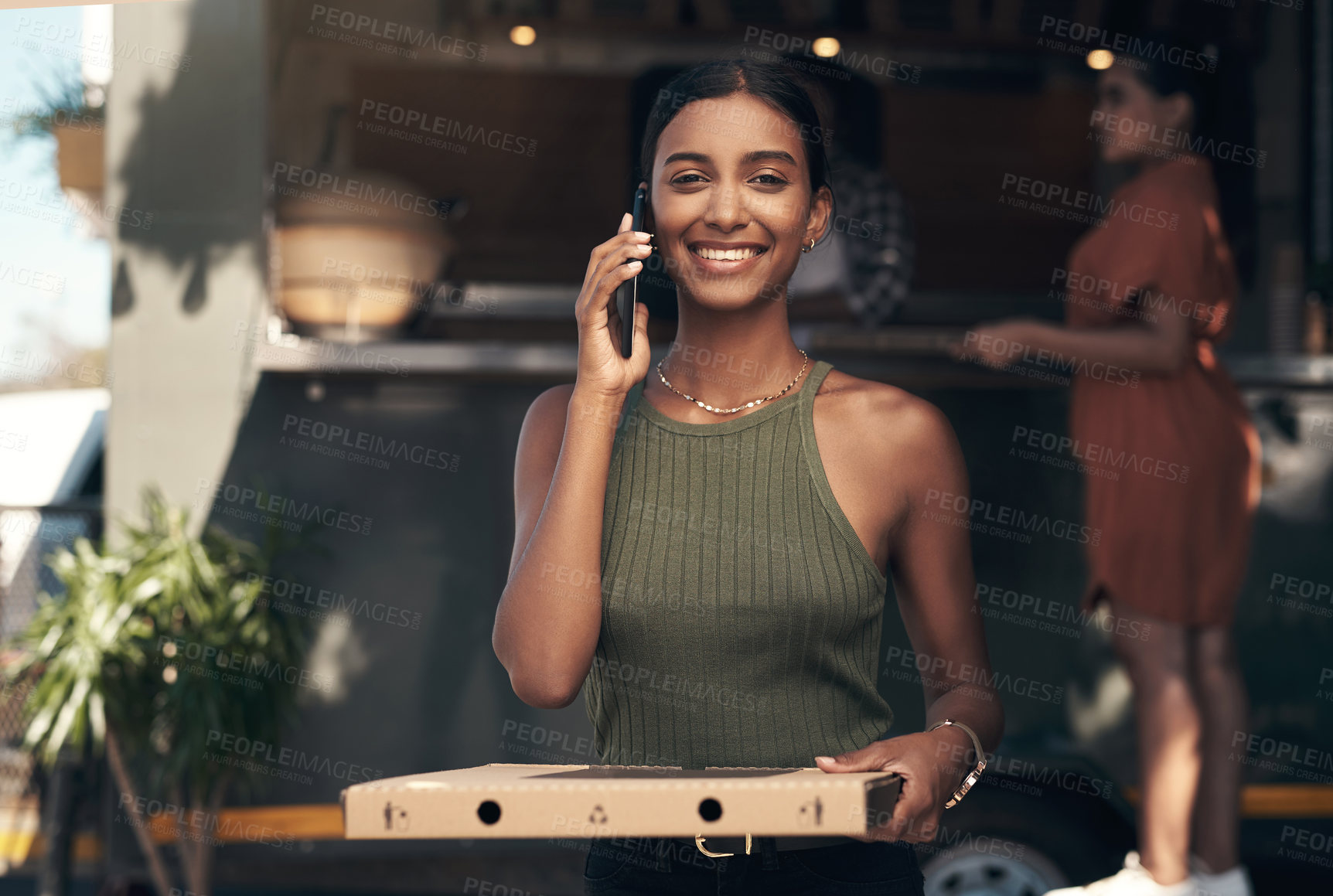 Buy stock photo Shot of an attractive young woman standing outside a restaurant and using her cellphone while holding her pizza