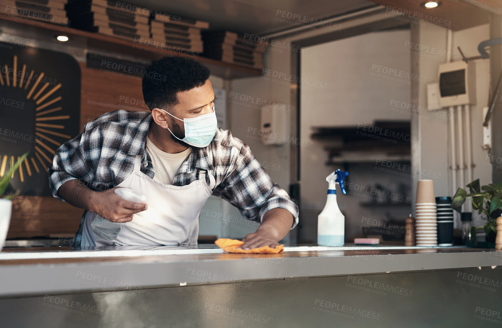 Buy stock photo Shot of a young man standing alone and sanitising the counter tops in his restaurant while wearing a face mask