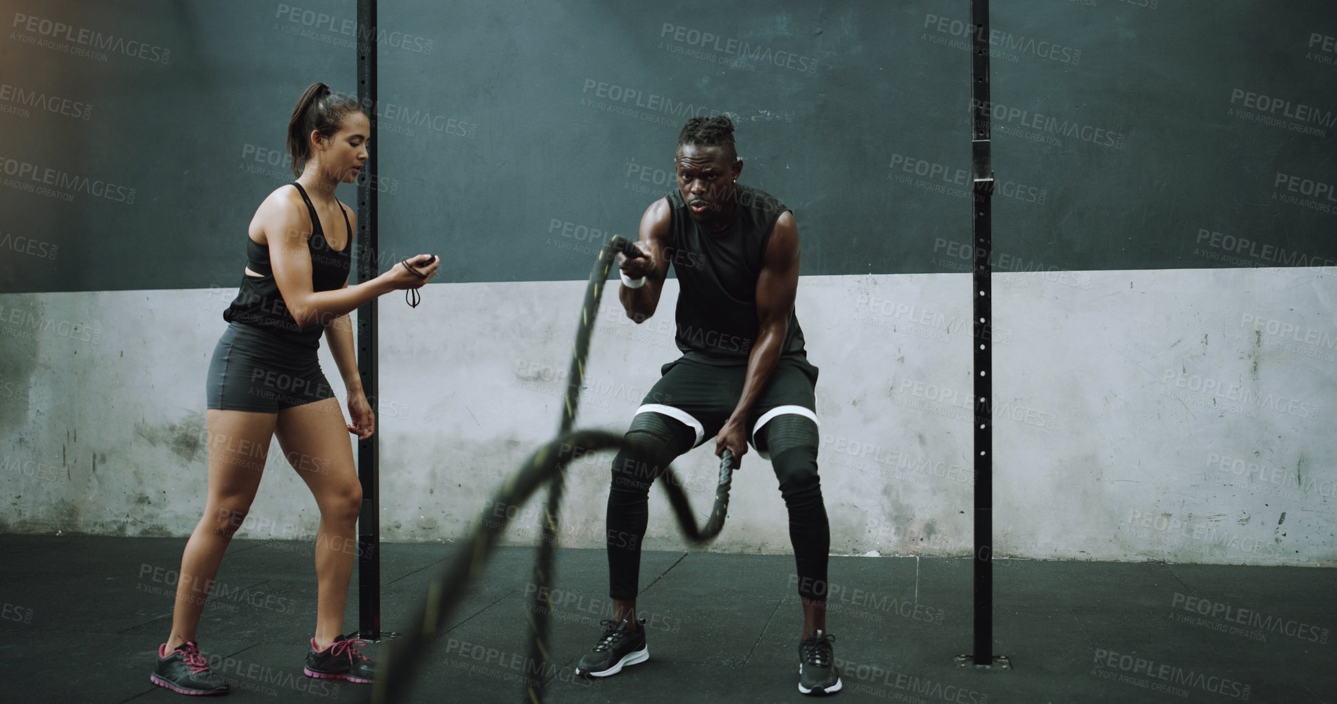 Buy stock photo Battle rope, exercise and man with personal trainer and stopwatch at gym for training goals on wall background. Heavy, full body and male with coach at sports center for power, workout or resilience