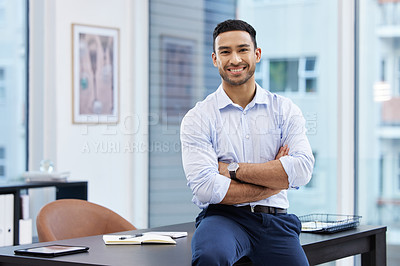 Buy stock photo Shot of a handsome young businessman sitting on his desk in the office with his arms folded