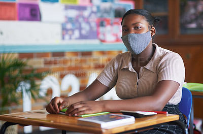 Buy stock photo Shot of a young girl sitting alone in her classroom at school and wearing a face mask